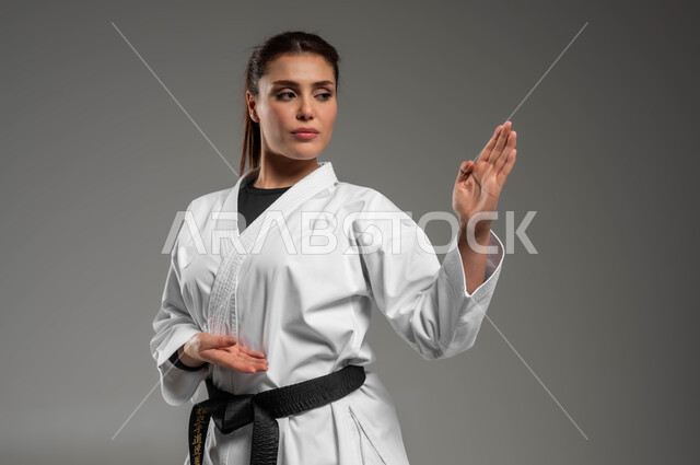أنشطة ورياضات تنافسية، تطبيق خطوات الكاتا، تنفيذ تمارين رياضية للمحافظة على اللياقة، فن الدفاع عن النفس, تعلم فنون قتال يابانية، بورتريه مقرب لفتاة عربية خليجية سعودية ترتدي الزي الرياضي الأبيض مع حزام أسود تمارس الكاراتيه بتعابير التركيز، خلفية رمادية