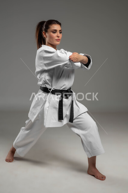 القيام بحركات قتالية خاصة وفن الدفاع عن النفس, تعلم فنون قتال يابانية وتطبيق خطوات الكاتا، بورتريه  لفتاة عربية خليجية سعودية ترتدي الزي الرياضي الأبيض مع حزام أسود تمارس الكاراتيه بإيماءات التركيز والانتباه، رياضات و تمارين نسائية، صورة لكامل الجسم، خلفية رمادية