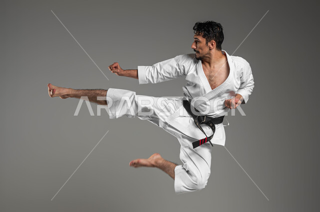 ممارسة فنون قتال يابانية بإيماءات التركيز و الانتباه، مفهوم اللياقة البدنية، تطبيق خطوات الكاتا، التدرب على فن الدفاع عن النفس, بورتريه لرجل عربي خليجي سعودي يرتدي الزي الرياضي الأبيض مع حزام أسود يمارس الكاراتيه، صورة لكامل الجسم، خلفية رمادية