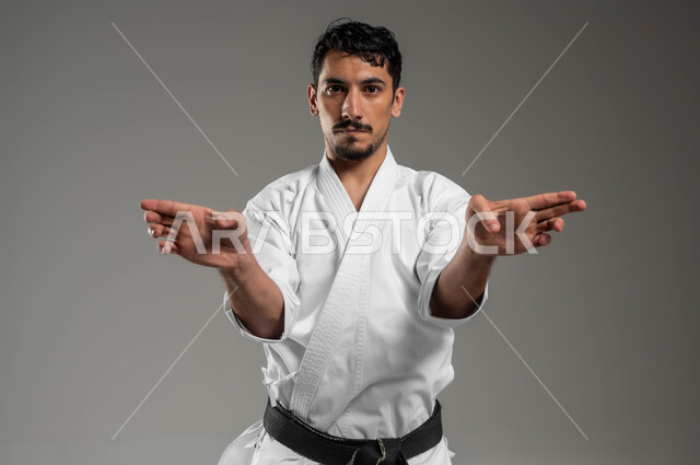 ممارسة فنون قتال يابانية بإيماءات التركيز و الانتباه، مفهوم اللياقة البدنية، تطبيق خطوات الكاتا، التدرب على حركات قتالية خاصة وفن الدفاع عن النفس, بورتريه مقرب لرجل عربي خليجي سعودي يرتدي الزي الرياضي الأبيض مع حزام أسود يمارس الكاراتيه، خلفية رمادية
