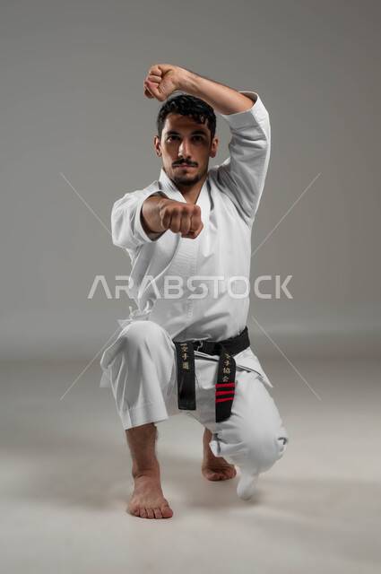 Looking at the camera with confidence, Japanese martial arts, applying kata steps, practicing special combat movements and the art of self-defense, portrait of a Saudi Arabian Gulf man wearing a white sports uniform with a black belt practicing karate with gestures of focus and attention, full body photo, gray background