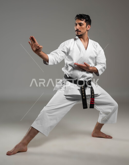 تطبيق خطوات الكاتا، التدرب على حركات قتالية خاصة وفن الدفاع عن النفس, بورتريه  لرجل عربي خليجي سعودي يرتدي الزي الرياضي الأبيض مع حزام أسود يمارس الكاراتيه بإيماءات التركيز والانتباه، فنون قتال يابانية، مفهوم اللياقة البدنية، صورة لكامل الجسم، خلفية رمادية