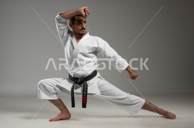 التدرب على حركات قتالية خاصة وفن الدفاع عن النفس, بورتريه  لرجل عربي خليجي سعودي يرتدي الزي الرياضي الأبيض مع حزام أسود يمارس الكاراتيه بإيماءات التركيز والانتباه، فنون قتال يابانية، تطبيق خطوات الكاتا، مفهوم اللياقة البدنية، صورة لكامل الجسم، خلفية رمادية