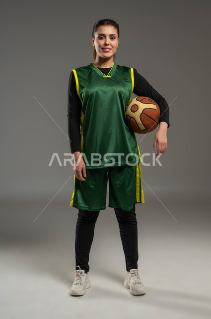 A healthy lifestyle, doing sports exercises, training to master playing basketball, maintaining physical fitness, recreational activities and women’s sports, a portrait of a smiling Saudi Gulf Arab player wearing sports clothes, holding a ball in her hand, full body photo, gray background
