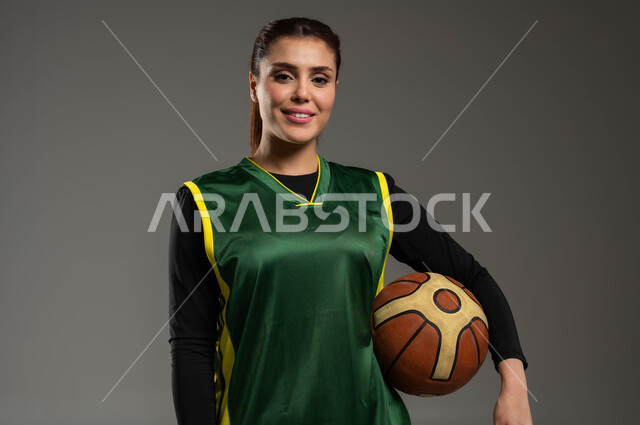 النظر إلى الكاميرا بإيماءات الثقة، نمط حياة صحي، إجراء تمرينات رياضية، التدرب على إتقان لعب كرة السلة، المحافظة على اللياقة البدنية، أنشطة ترفيهية ورياضات نسائية، بورتريه مقرب للاعبة عربية خليجية سعودية مبتسمة ترتدي ملابس رياضية تحمل بيدها الكرة، خلفية رمادية