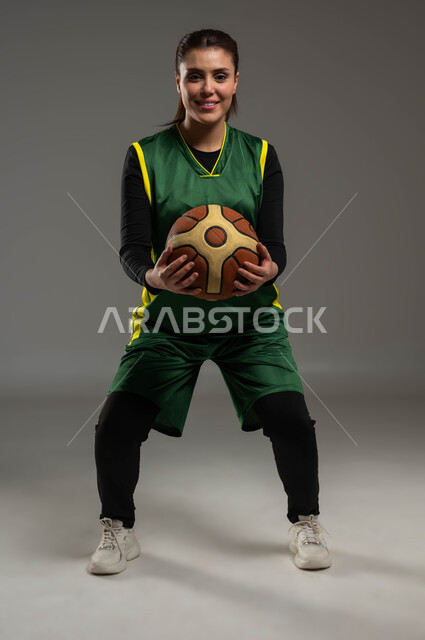 الاستعداد لتصويب الهدف، إجراء تمرينات رياضية، التدرب على إتقان لعب كرة السلة، المحافظة على اللياقة البدنية، أنشطة ترفيهية ورياضات نسائية، بورتريه للاعبة عربية خليجية سعودية مبتسمة ترتدي ملابس رياضية، أسلوب الحياة الصحية، صورة لكامل الجسم، خلفية رمادية