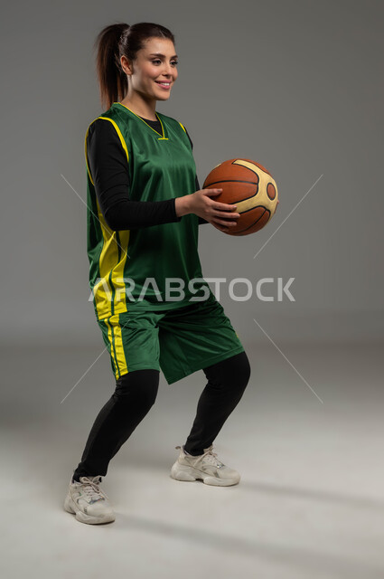 الاستعداد لرمي الكرة، إجراء تمرينات رياضية، التدرب على إتقان لعب كرة السلة، المحافظة على اللياقة البدنية، أنشطة ترفيهية ورياضات نسائية، بورتريه للاعبة عربية خليجية سعودية مبتسمة ترتدي ملابس رياضية، أسلوب الحياة الصحية، صورة لكامل الجسم، خلفية رمادية
