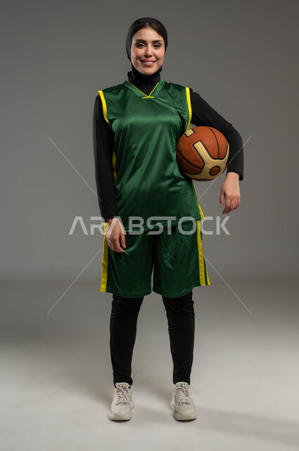 النظر الى الكاميرا بإيماءات الثقة، التدرب على إتقان لعب كرة السلة، المحافظة على اللياقة البدنية، أنشطة ترفيهية ورياضات نسائية، بورتريه للاعبة عربية خليجية سعودية محجبة ترتدي ملابس رياضية تمسك بيدها الكرة، أسلوب الحياة الصحية، صورة لكامل الجسم، خلفية رمادية