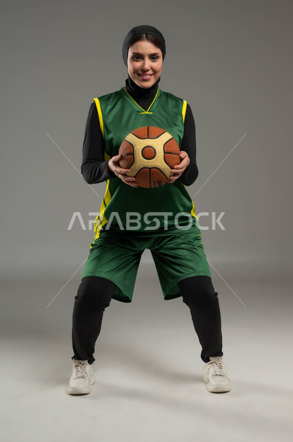 الاستعداد لرمي الكرة، التدرب على إتقان لعب كرة السلة، النظر بإيماءات السعادة والسرور، المحافظة على اللياقة البدنية، أنشطة ترفيهية ورياضات نسائية، بورتريه للاعبة عربية خليجية سعودية محجبة ترتدي ملابس رياضية، أسلوب الحياة الصحية، صورة لكامل الجسم، خلفية رمادية