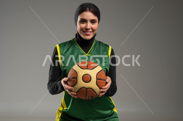 التدرب على إتقان لعب كرة السلة، الوقوف باستقامة والنظر بإيماءات السعادة والسرور، المحافظة على اللياقة البدنية، أنشطة ترفيهية ورياضات نسائية، بورتريه مقرب للاعبة عربية خليجية سعودية محجبة ترتدي ملابس رياضية تمسك بيدها الكرة، خلفية رمادية