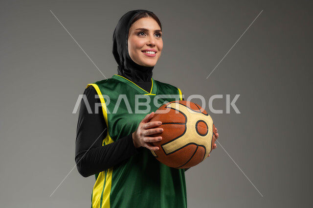 النظر لمكان ما بإيماءات السعادة والسرور، المحافظة على اللياقة البدنية، أسلوب الحياة الصحية، أنشطة ترفيهية ورياضات نسائية، التدرب على إتقان لعب كرة السلة، بورتريه مقرب للاعبة عربية خليجية سعودية محجبة ترتدي ملابس رياضية تمسك بيدها الكرة، خلفية رمادية