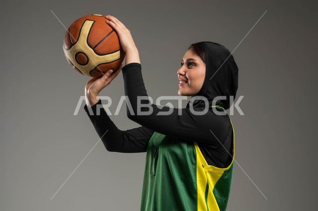Women's entertainment and sports activities, healthy lifestyle, training to master basketball, maintaining physical fitness, close-up portrait from the side of a veiled Saudi Gulf Arab player wearing green sports clothing preparing to throw the ball, gestures and expressions of focus and attention, gray background