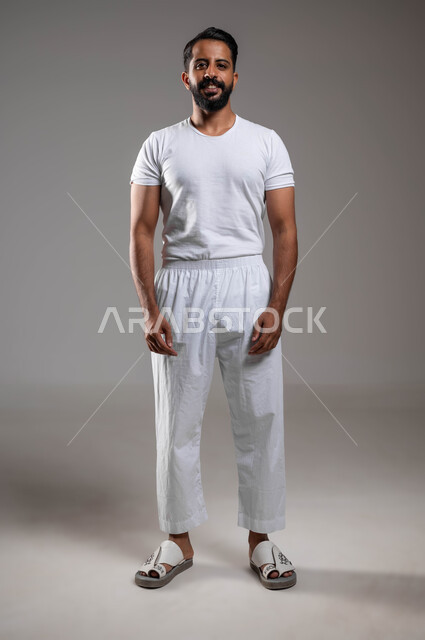 ملابس داخلية رجالية مريحة باللون الابيض، إيماءات السعادة والسرور، الوقوف باستقامة والنظر إلى الكاميرا، أزياء عربية سعودية، مفهوم لغة الجسد، بورتريه لرجل عربي خليجي سعودي وسيم  يرتدي سروال وفنيله بيضاء ينظر بفرح، صورة لكامل الجسم، خلفية رمادية