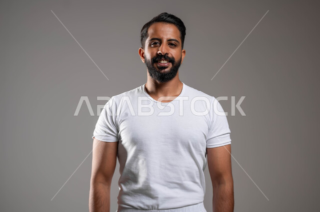 إيماءات السعادة والسرور، الوقوف باستقامة والنظر إلى الكاميرا، أزياء عربية سعودية، مفهوم لغة الجسد، بورتريه مقرب لرجل عربي خليجي سعودي وسيم  يرتدي سروال وفنيله بيضاء ينظر بفرح، ملابس داخلية رجالية مريحة باللون الابيض، خلفية رمادية