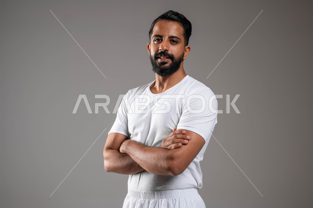 إيماءات الثقة بالنفس، الوقوف باستقامة والنظر إلى الكاميرا، مفهوم لغة الجسد، بورتريه مقرب لرجل عربي خليجي سعودي وسيم  يرتدي سروال وفنيله بيضاء يقف بايدي متقاطعة ، ملابس داخلية رجالية مريحة باللون الابيض، خلفية رمادية