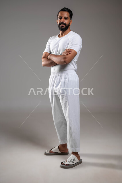 الوقوف باستقامة والنظر إلى الكاميرا، إيماءات الثقة بالنفس، مفهوم لغة الجسد، بورتريه لرجل عربي خليجي سعودي وسيم  يرتدي سروال وفنيله بيضاء يقف بايدي متقاطعة ، ملابس داخلية رجالية مريحة باللون الابيض، صورة لكامل الجسم، خلفية رمادية