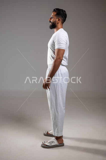 ملابس داخلية رجالية مريحة باللون الابيض، إيماءات السعادة والسرور، الوقوف باستقامة، أزياء عربية سعودية، مفهوم لغة الجسد، بورتريه من الجانب لرجل عربي خليجي سعودي وسيم  يرتدي سروال وفنيله بيضاء ينظر بثقة، صورة لكامل الجسم، خلفية رمادية