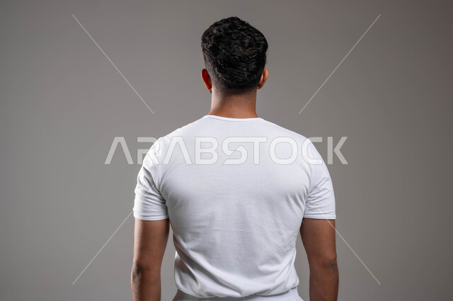 الاعتراض عن شيء ما، إيماءات تدل على الرفض، أزياء وملابس عربية سعودية، ملابس داخلية رجالية مريحة باللون الابيض، مفهوم لغة الجسد، بورتريه مقرب من الخلف لرجل عربي خليجي سعودي وسيم  يرتدي سروال وفنيله بيضاء يقف باستقامة، خلفية رمادية