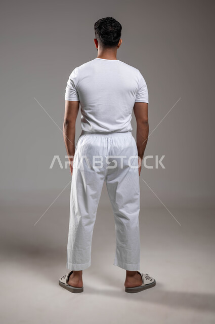 إيماءات تدل على الرفض، أزياء وملابس عربية سعودية، الاعتراض عن شيء ما، ملابس داخلية رجالية مريحة باللون الابيض، مفهوم لغة الجسد، بورتريه من الخلف لرجل عربي خليجي سعودي وسيم  يرتدي سروال وفنيله بيضاءيقف باستقامة، صورة لكامل الجسم، خلفية رمادية