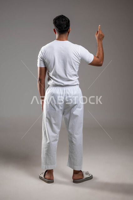الوقوف باستقامة و الإشارة للأعلى، التوجه و الإشارة لشيء ما، مفهوم لغة الجسد، بورتريه  من الخلف لرجل عربي خليجي سعودي وسيم  يرتدي سروال ونفيلة بيضاء يشير باصبعه السبابة إلى مكان ما، ملابس داخلية رجالية مريحة باللون الابيض، صورة لكامل الجسم، خلفية رمادية