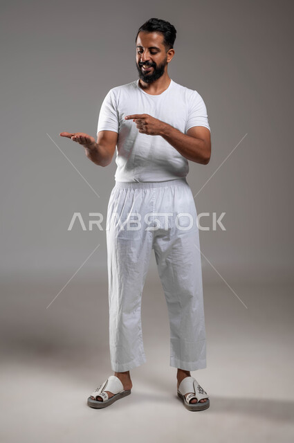 النظر إلى اليدين بإيماءات السعادة والسرور، التوجه و الإشارة لشيء ما، مفهوم لغة الجسد، بورتريه لرجل عربي خليجي سعودي وسيم مبتسم يرتدي سروال ونفيلة بيضاء يشير باصبعه السبابة إلى يده الأخرى، ملابس داخلية رجالية مريحة باللون الابيض، صورة لكامل الجسم، خلفية رمادية
