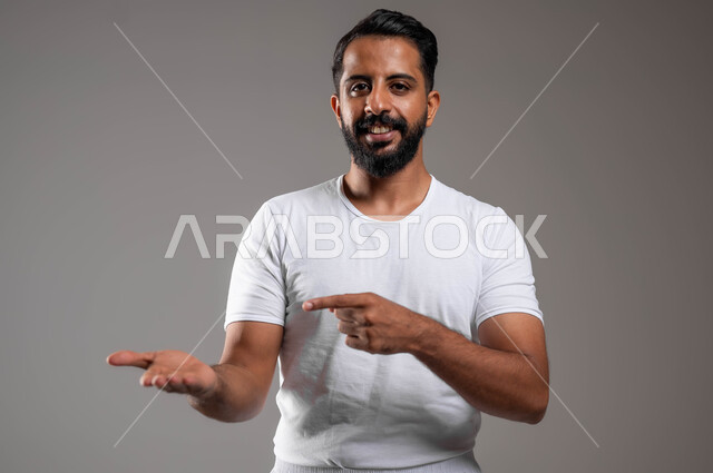 النظر إلى الكاميرا بإيماءات السعادة والسرور، التوجه و الإشارة لشيء ما، مفهوم لغة الجسد، بورتريه مقرب لرجل عربي خليجي سعودي وسيم مبتسم يرتدي سروال ونفيلة بيضاء يشير باصبعه السبابة إلى يده الأخرى، ملابس داخلية رجالية مريحة باللون الابيض، خلفية رمادية