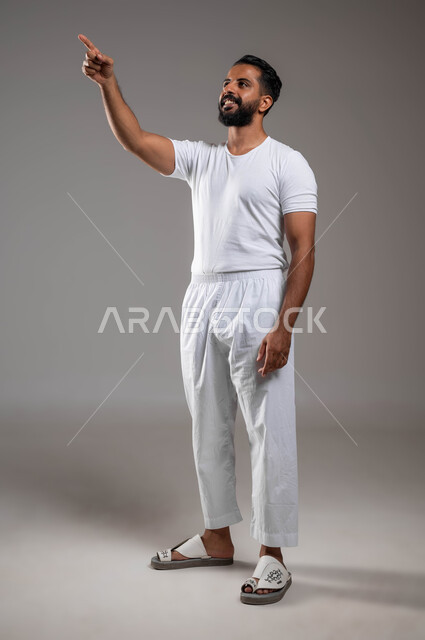 مفهوم لغة الجسد، التوجه و الإشارة لشيء ما، بورتريه لرجل عربي خليجي سعودي وسيم مبتسم يرتدي سروال ونفيلة بيضاء يشير باصبعه السبابة إلى شيء، ملابس داخلية رجالية مريحة باللون الابيض، إيماءات السعادة والسرور، صورة الجسم بالطول الكامل، خلفية رمادية