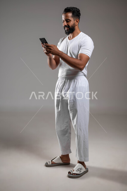 Communicating with family and friends, shopping online, browsing mobile programs and applications, a portrait of a Saudi Gulf Arab man wearing pants and a white T-shirt holding a mobile phone in his hand, using advanced devices and technologies, a full-body portrait, gray background