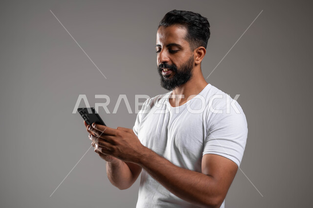 تصفح برامج و تطبيقات الجوال، التواصل مع الأهل والأصدقاء، التسوق عبر الانترنت، بورتريه  مقرب لرجل عربي سعودي خليجي يرتدي السروال والفنيله البيضاء يحمل بيده الهاتف المحمول ، استخدام الأجهزة والتقنيات المتطورة، خلفية رمادية