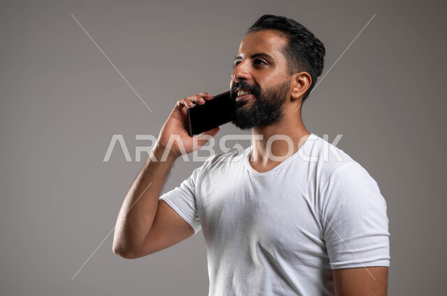 Communicating with family and friends, making phone calls via mobile phone, a close-up portrait of a smiling Saudi Arabian Gulf man wearing pants and a white T-shirt holding a mobile phone in his hand, the concept of development and modern technology, the use of advanced devices and technologies, gray background