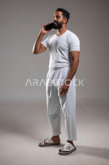 Making phone calls via mobile phone, portrait of a smiling Saudi Arabian Gulf man wearing pants and a white T-shirt holding a mobile phone in his hand, using advanced devices and technologies, communicating with family and friends, full-length body photo, gray background