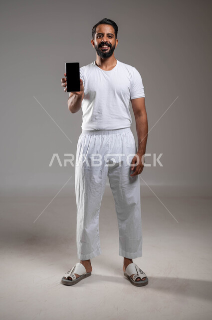 Looking at the camera with gestures of happiness and pleasure, the concept of development and technology, the use of modern and advanced technologies, a portrait of a smiling Saudi Arabian Gulf man wearing pants and a T-shirt, standing straight, holding a mobile phone in his hand, showing the mobile screen, full-body portrait, gray background