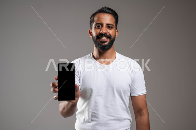 مفهوم التطور والتكنولوجيا، النظر إلى الكاميرا بإيماءات السعادة والسرور، استخدام التقنيات الحديثة والمتطورة، بورتريه مقرب لرجل عربي خليجي سعودي مبتسم برتدي سروال وفنيله يقف باستقامة يحمل بيده الهاتف المحمول يقوم بعرض شاشة الموبايل، خلفية رمادية
