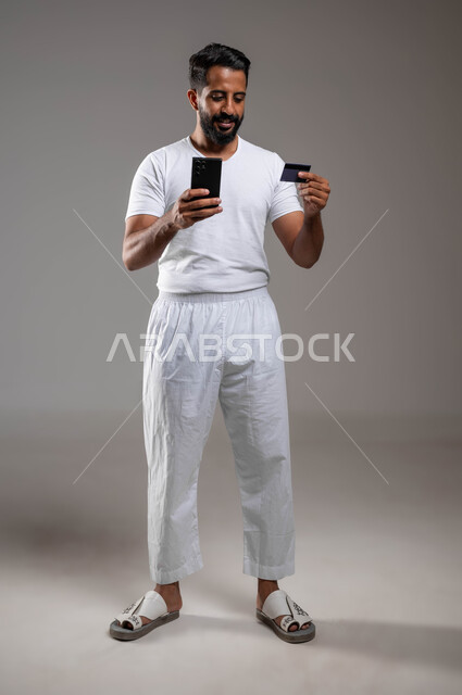 الشراء والدفع من خلال برامج وتطبيقات الجوال، التقنيات الحديثة والمتطورة، بورتريه لرجل عربي خليجي سعودي مبتسم برتدي سروال وفنيله بيضاء يمسك بيده الهاتف المحمول وبطاقة الإئتمان البنكية، تسديد الفواتير بالبطاقات الإلكترونية، صورة لكامل الجسم، خلفية رمادية