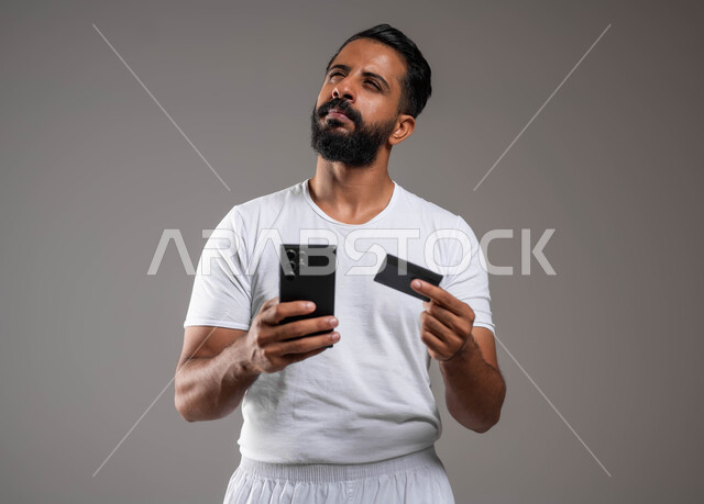 التفكير بعمق وحيرة، الشراء والدفع من خلال برامج وتطبيقات الجوال، بورتريه مقرب لرجل عربي خليجي سعودي برتدي سروال وفنيله يجلس على كرسي يحمل بيده الهاتف المحمل وبطاقة الإئتمان البنكية، تسديد الفواتير بالبطاقات الإلكترونية، استخدام خلفية رمادية