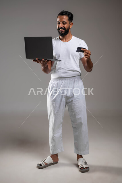 The possibility of paying bills electronically through programs, paying through laptop applications, using an electronic bank card, a portrait of a smiling Saudi Arabian Gulf man wearing pants and a white T-shirt, standing holding a laptop, full-length body portrait, gray background