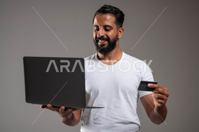 استخدام البطاقة البنكية الالكترونية، إيماءات السعادة والسرور، إمكانية تسديد الفواتير عبر البرامج الكترونيا، الدفع عبر تطبيقات اللابتوب، بورتريه مقرب لرجل عربي سعودي خليجي مبتسم يرتدي السروال والفنيله البيضاء يقف ممسكا جهاز الحاسوب المحمول، خلفية رمادية