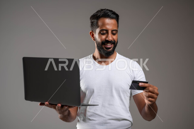 إيماءات السعادة والسرور، إمكانية تسديد الفواتير عبر البرامج الكترونيا، الدفع عبر تطبيقات اللابتوب، استخدام البطاقة البنكية الالكترونية، بورتريه مقرب لرجل عربي سعودي خليجي مبتسم يرتدي السروال والفنيله البيضاء يقف ممسكا جهاز الحاسوب المحمول، خلفية رمادية