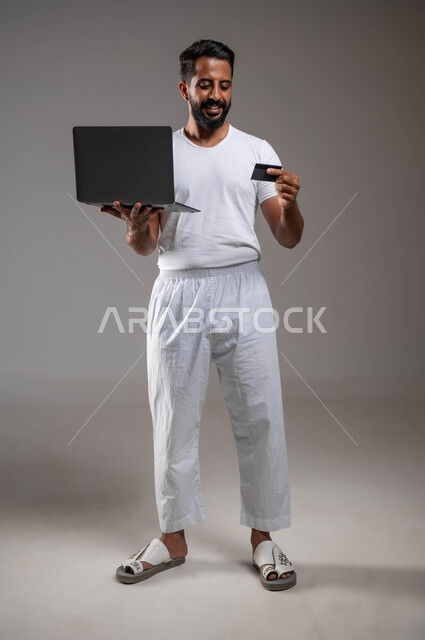 الدفع عبر تطبيقات اللابتوب، استخدام البطاقة البنكية الالكترونية، بورتريه لرجل عربي سعودي خليجي مبتسم يرتدي السروال والفنيله البيضاء يقف ممسكا جهاز الحاسوب المحمول، إمكانية تسديد الفواتير عبر البرامج الكترونيا، صورة الجسم بالطول الكامل، خلفية رمادية
