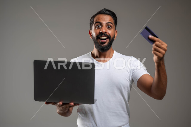 استخدام البطاقة البنكية الالكترونية، مفهوم الدفع والتسديد عن بعد عبر تطبيقات اللابتوب، بورتريه مقرب لشاب عربي سعودي خليجي يرتدري السروال والفنيلة البيضاء, ايماءات الحماس والفرح, خلفية رمادية