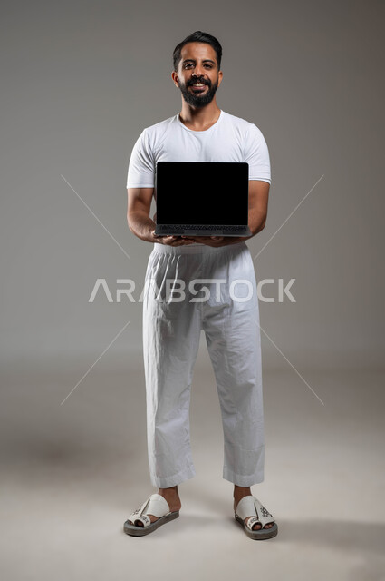 استخدام الأجهزة التقنية المتطورة, دور التقنيات الحديثة في عملية البحث عن المعلومات المفيدة, بورتريه لشاب عربي سعودي خليجي يرتدي السروال والفنيلة البيضاء يمسك بيده الحاسب المحمول بشاشة سوداء فارغة, ايماءات الفرح والحماس, صورة الجسم بالطول الكامل, خلفية رمادية