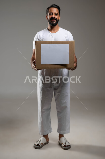 Organisations caritatives et aide humanitaire, œuvre caritative islamique en Arabie Saoudite, portrait d'un jeune homme saoudien du Golfe portant un pantalon et un T-shirt tenant une boîte de dons à la main, initiatives et distribution de fournitures aux nécessiteux, photo du corps en pied, fond gris