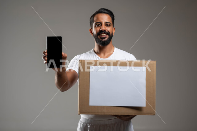 Projets et organisations caritatives en ligne, applications électroniques pour collecter des dons, portrait en gros plan d'un jeune homme arabe du Golfe saoudien portant un pantalon et un T-shirt blanc tenant une boîte de dons, affichant le téléphone portable avec un écran noir vierge, fond gris