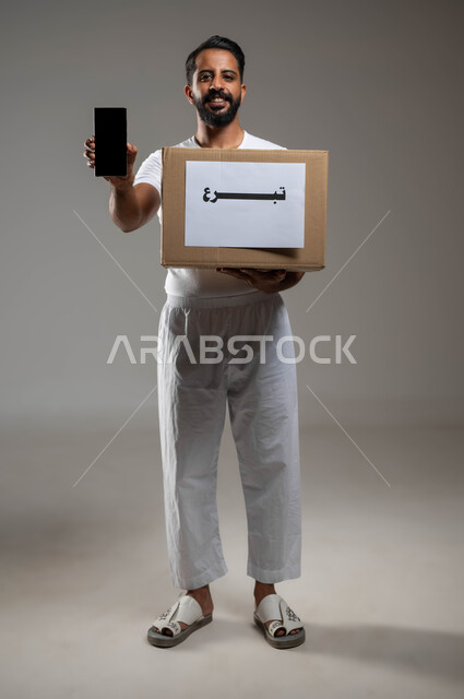 التطبيقات الالكترونية لجمع التبرعات, مشاريع وجمعيات خيرية عبر الأونلاين, بورتريه لشاب عربي خليجي سعودي يرتدي السروال والفنيلة البيضاء يحمل صندوق التبرع, عرض الهاتف المحمول بشاشة سوداء فارغة, صورة الجسم بالطول الكامل, خلفية رمادية