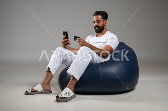 التسوق الالكتروني عبر الانترنت, استخدام البطاقة البنكية, بورتريه لشاب عربي سعودي خليجي يرتدي السروال والفنيلة البيضاء يجلس في مكان مريح يمسك بيده الهاتف المحمول وبطاقة البنك, خلفية رمادية