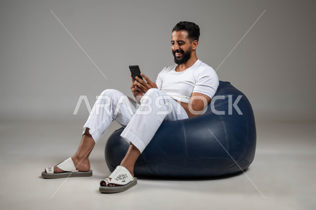 تطور التكنولوجيا والأجهزة التقنية، إجراء مكالمة فيديو مع الأهل والأصدقاء، بورتريه لشاب عربي سعودي خليجي مبتسم يرتدي السروال والفنيله البيضاء يجلس في مكان مريح يحمل الهاتف المحمول،  خلفية رمادية