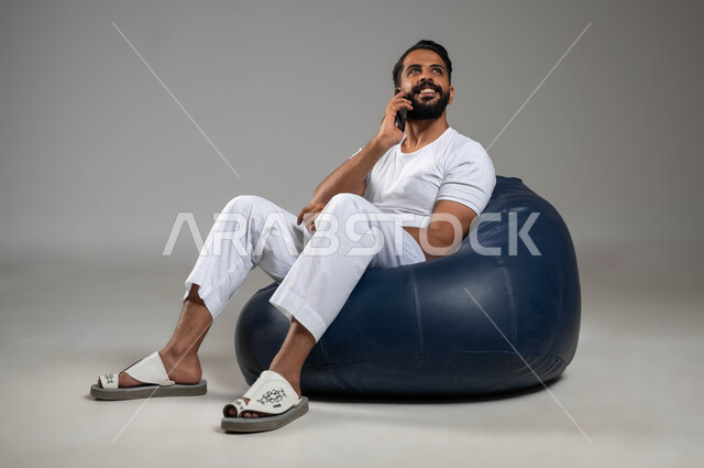 تطور التكنولوجيا والأجهزة التقنية, اجراء مكالمة هاتفية مع الاهل والاصدقاء عبر الجوال, ايماءات الفرح والسعادة, بورتريه لشاب عربي سعودي خليجي يرتدري السروال والفنيلة البيضاء يجلس في مكان مريح, خلفية رمادية