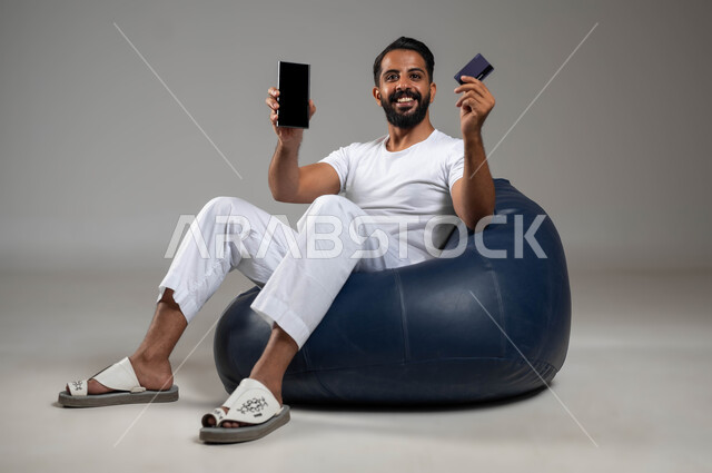 Assis joyeusement et joyeusement, utilisant une carte bancaire pour le paiement électronique, un portrait d'un homme souriant du Golfe saoudien portant un pantalon et un T-shirt blanc, affichant le téléphone portable avec un écran noir vierge, la possibilité de payer des factures via des programmes électroniques, photo du corps entier, fond gris