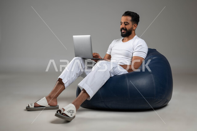 إدارة الأعمال عبر الأونلاين, مفهوم استخدام الأجهزة والتقنيات الحديثة والمتطورة, بورتريه لشاب عربي خليجي سعودي يرتدي السروال والفنيلة البيضاء يستخدم الحاسب المحمول, صورة الجسم بالكامل, خلفية رمادية