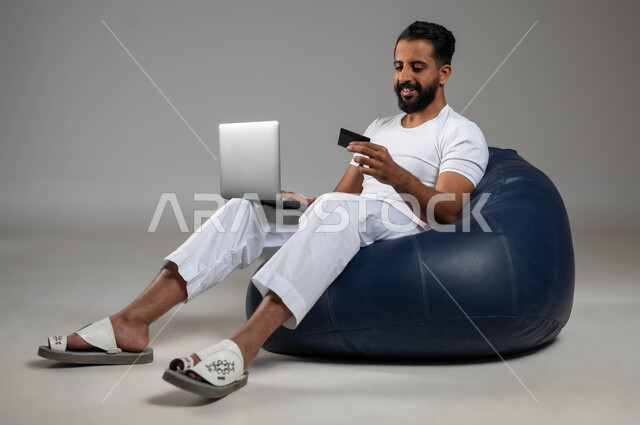 إمكانية تسديد الفواتير عبر البرامج الالكترونيه, استخدام البطاقة البنكية للدفع الالكتروني، بورتريه لرجل عربي سعودي خليجي مبتسم يرتدي السروال والفنيله البيضاء يجلس في مكان مريح يعمل على جهاز الحاسب المحمول، صورة الجسم بالكامل، خلفية رمادية